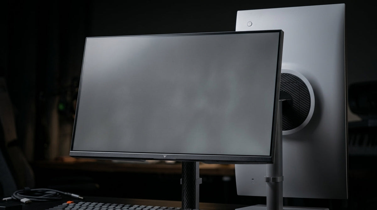 A technical close-up of a high-end monitor screen in a dark creative workspace, displaying a gray test pattern that reveals subtle clouding and Mura effects.